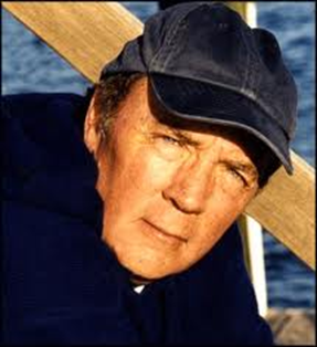 A close-up portrait of a middle-aged man wearing a black baseball cap. He appears to be in his late 60s or early 70s with short dark hair and wrinkles on his face. He is looking directly at the camera with a serious expression. The background is blurred but it seems to be a body of water possibly a lake or ocean. The man is wearing a dark blue jacket and there is a wooden railing visible in the background. The overall mood of the image is somber and contemplative.