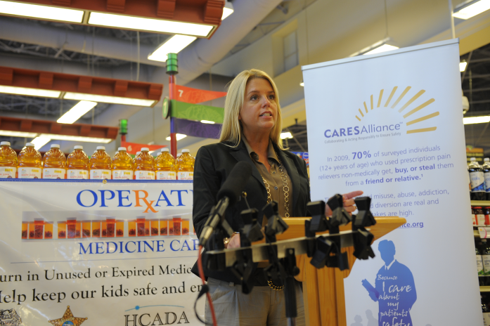 A woman standing behind a podium in a store. She is speaking into a microphone and appears to be giving a presentation. Behind her there is a banner that reads "Operate Medicine Care" and a sign that says "Turn in Unused or Expired Medicine Help keep our kids safe and secure". There are shelves with various items in the background. The woman is wearing a black blazer and has blonde hair.