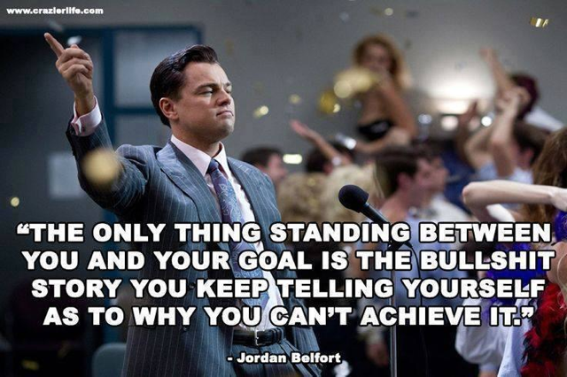 A man in a suit pointing upwards with a quote from Jordan Belfort that reads "The only thing standing between you and your goal is the bullshit story you keep telling yourself as to why you can't achieve it." The man is standing in front of a microphone and appears to be giving a speech or presentation. Behind him there is a crowd of people with their hands in the air cheering and celebrating. The background is blurred but it seems like the focus is on the man and the quote.