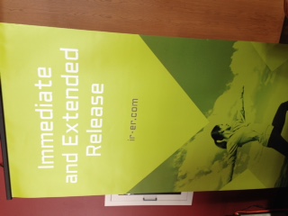 A photograph of a book titled "Immediate and Extended Release". The book is lying on a wooden surface and is open to a page with a green background. The title of the book is written in white text on the left side of the page. On the right side there is an image of a person holding a camera and taking a photo. The person is wearing a black shirt and jeans and is standing in front of a mountain landscape. The image appears to be taken in a room with a red wall in the background.