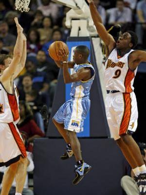 A basketball game in progress. Two players from the opposing team wearing orange and white jerseys are in the air attempting to block a shot by one of the players who is wearing a blue jersey with the number 11 on it. The player in the blue jersey is holding the ball and is in mid-air with his arms stretched out to the sides. The other player wearing the orange jersey is jumping up to try and block the shot. The background shows a crowd of spectators in the stands. The image appears to have been taken from the sidelines of the court.