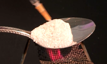 A silver spoon with a wooden handle resting on top of a wire mesh strainer. The strainer is filled with a white substance which appears to be sugar or flour. The background is black making the spoon and strainer stand out. The spoon is positioned in the center of the image with the handle on the left side and the strainer on the right side. The image is taken from a slightly elevated angle looking down on the spoon.