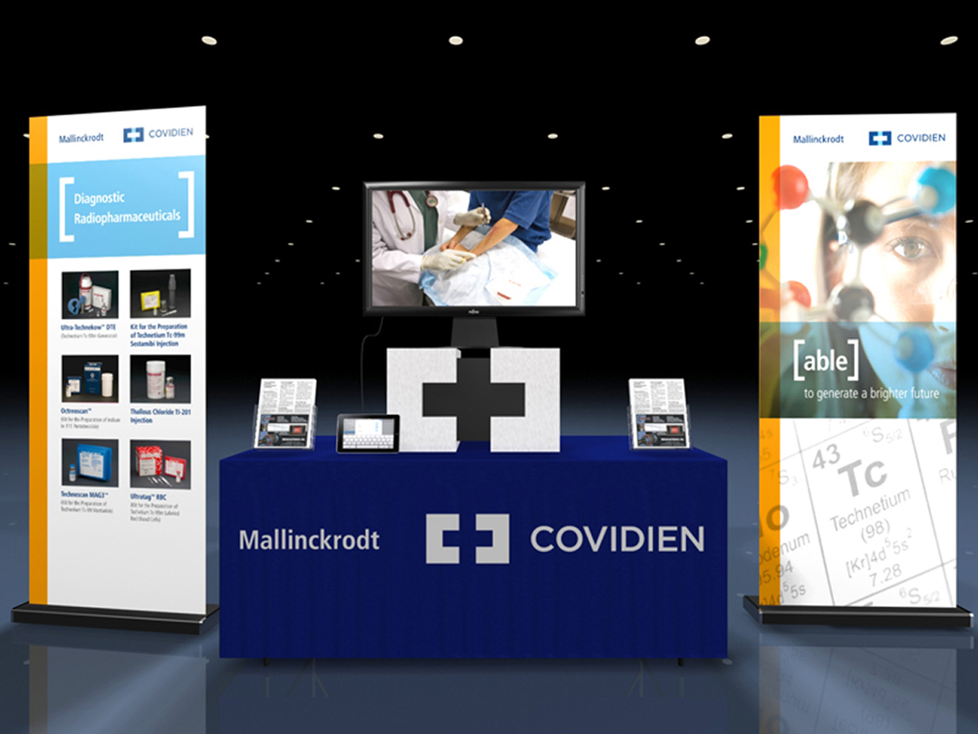 A trade show booth with a blue table in the center. On the table there is a large screen displaying a medical image of a patient lying on a bed with a stethoscope around their neck. Next to the screen there are two banners with the text "Covidien" and "Table" on them. The banners also have images of medical equipment and information about the company. The booth is set up in a dark room with a black background.