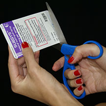 A pair of hands holding a purple and white card with a red and white label on it. The card appears to be a medical ID card as indicated by the text on the label. The person's hands are holding the card with one hand and the other hand is holding a blue pair of scissors. The scissors are positioned in a way that the person is using them to cut through the card. The background is black making the blue scissors and the card stand out.