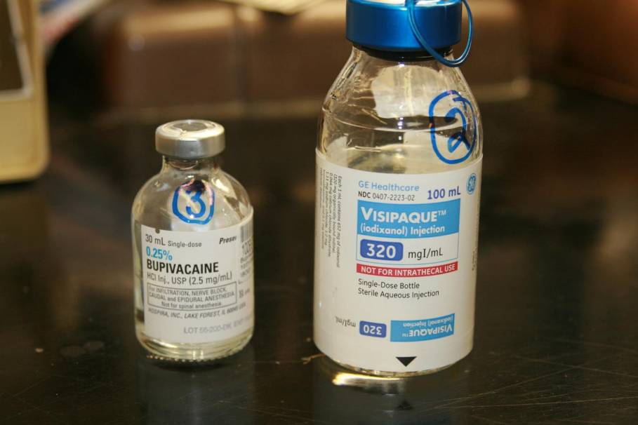 Two vials of vaccine on a black countertop. The vials are made of clear plastic and have a blue cap. The label on the vials is white with blue text and a blue logo. The text on the label reads "Visipaque" and "320 mg/ml". The label also mentions that the vial contains 100 ml of the vaccine. The background is blurred but it appears to be a laboratory setting with other medical equipment visible.