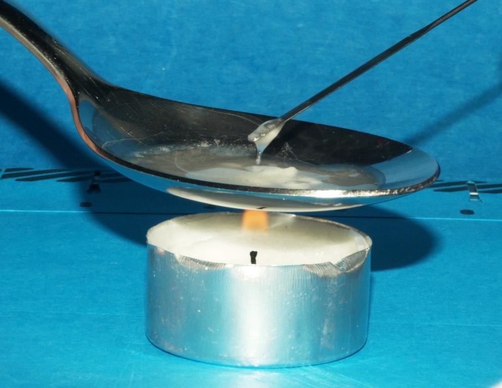 A close-up of a spoon holding an unidentified substance being held over a small white votive candle (with a silver wrapper). The candle is lit presumably to cook the substance. A metal rod - similar to a drink stirrer - is dipping into the substance to demonstrate the consistency of the substance. The background is light blue.