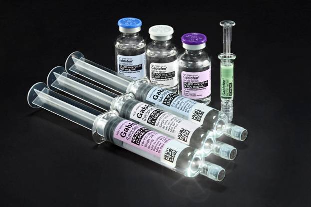 A group of six syringes of different sizes and shapes arranged in a row on a black background. There are six vials in total each with a different color - blue purple green and white. The vials are filled with a clear liquid and one of them has a label with a QR code on it. Next to the vials there is a small syringe with a green liquid inside. The syringe appears to be filled with the same liquid as the other vials. The background is completely black making the colors of the syringe and vials stand out.