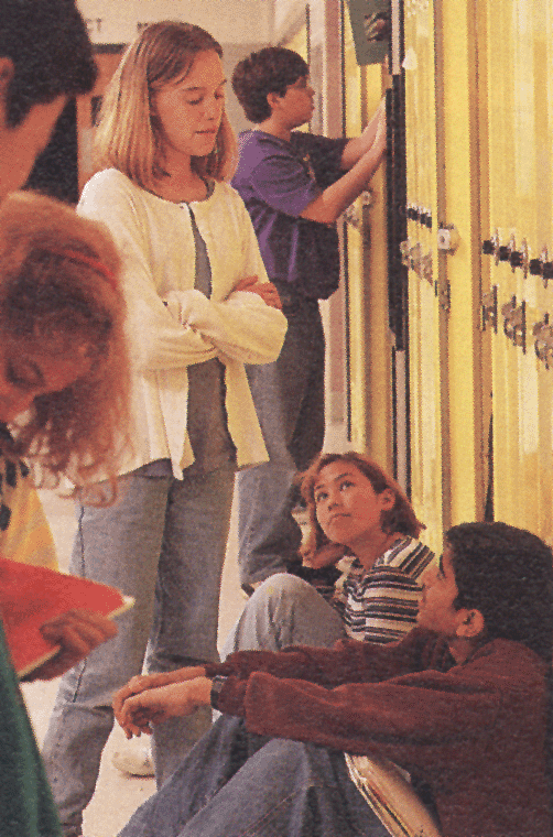 A group of young people in a hallway. There are four people in the image two boys and two girls standing in front of a row of yellow lockers. The girl in the center is standing with her arms crossed and appears to be explaining something to the other two boys. The boy on the left is holding a red book and is looking at the girl on the right who is sitting on the floor with a serious expression on her face. The other boy is standing next to the lockers and is also looking at something on the wall. It appears that the group is engaged in a discussion or activity.