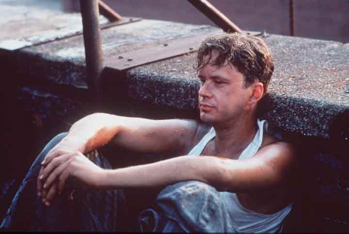 A young man sitting on a concrete bench. He is wearing a white tank top and blue jeans. He has short dark hair and is looking off to the side with a serious expression on his face. His arms are crossed over his chest and he appears to be deep in thought. The background is blurred but it seems to be an outdoor setting with a metal railing visible. The overall mood of the image is somber and contemplative.