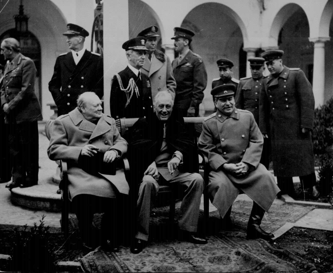 A black and white photograph of a group of men in military uniforms. The men are sitting on a bench in a courtyard with arches and pillars in the background. They are all wearing caps and jackets and some are standing while others are sitting.<br /><br />In the center of the image there is an older man sitting on the bench with a smile on his face. He is wearing a suit and tie and appears to be in his late 60s or early 70s. To his left there are two older men one wearing a coat and the other wearing a hat both of whom are looking at the camera. Behind them a few more men are standing and observing the group. The overall mood of the photograph is relaxed and casual.