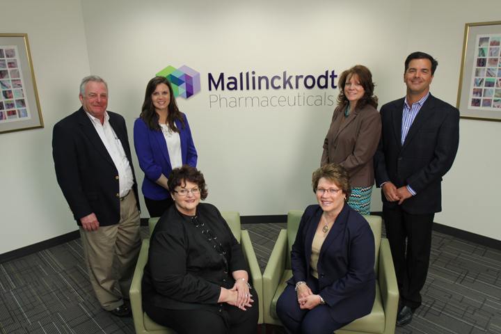 A group of six people standing in front of a wall with the logo of Mallinckrodt Pharmaceuticals. The logo is a colorful cube with the company name written in white letters. The people in the group are of different ages and ethnicities and they are all smiling and posing for the photo. On the left side of the image there is a man wearing a black suit and a woman wearing a blue blazer. Next to him there are two women sitting on green armchairs. The floor is covered with a gray carpet and there are several framed pictures hanging on the wall behind them.