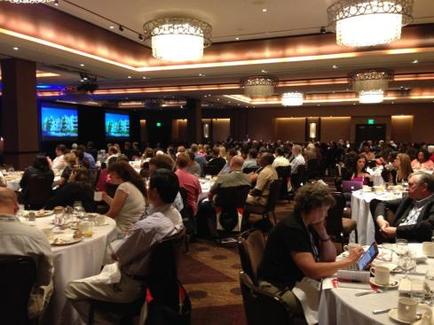 A large banquet hall with a high ceiling and chandeliers. The room is filled with people sitting at long tables facing a large screen on the left side of the room. The tables are covered with white tablecloths and are set with plates cups saucers and glasses. The people are engaged in conversation and appear to be engaged in a discussion. The walls are decorated with a patterned wallpaper and there is a large window on the right side. The overall atmosphere is formal and professional.