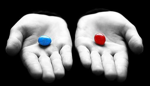 Two hands holding two pills - one blue and one red. The blue pill is on the left side of the image and the red pill is in the center. The hands are positioned in a way that the palms are cupped together with the fingers slightly spread apart. The background is black making the two pills stand out. The image is in black and white giving it a monochromatic tone.