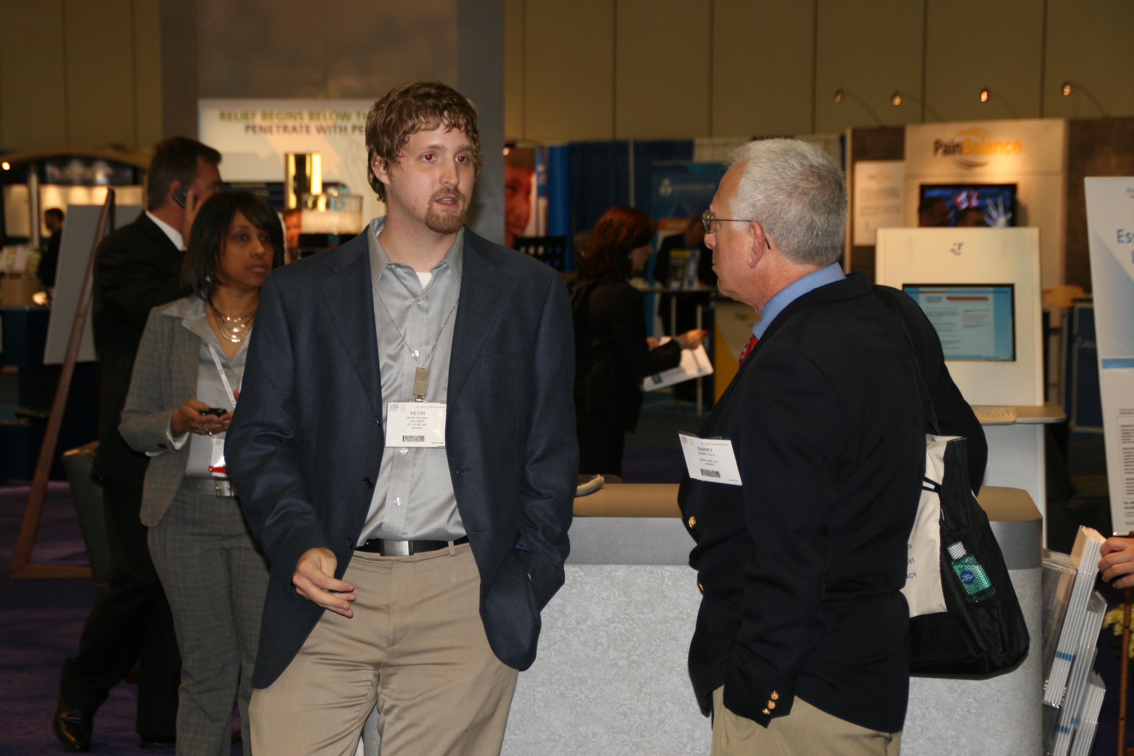 Two men engaged in a conversation at an exhibition. The man on the left is wearing a blue blazer and beige pants and has a name tag around his neck. He appears to be in his late twenties or early thirties and is looking off to the side with a serious expression on his face. On the right side of the image there is an older man wearing a black jacket and carrying a black bag. In the background there are other people walking around and a booth with a sign that reads "Exhibition". The booth is set up in a large room with a purple carpet and white walls.