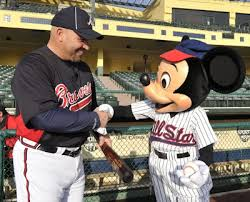 A baseball player from the Boston Red Sox team wearing a black jersey with the team's logo on it standing in a baseball stadium. He is holding a baseball bat and is shaking hands with a mascot Mickey Mouse who is wearing a red and white striped uniform and a baseball cap. The mascot is standing next to the player and appears to be congratulating him. The background shows the empty seats of the stadium.