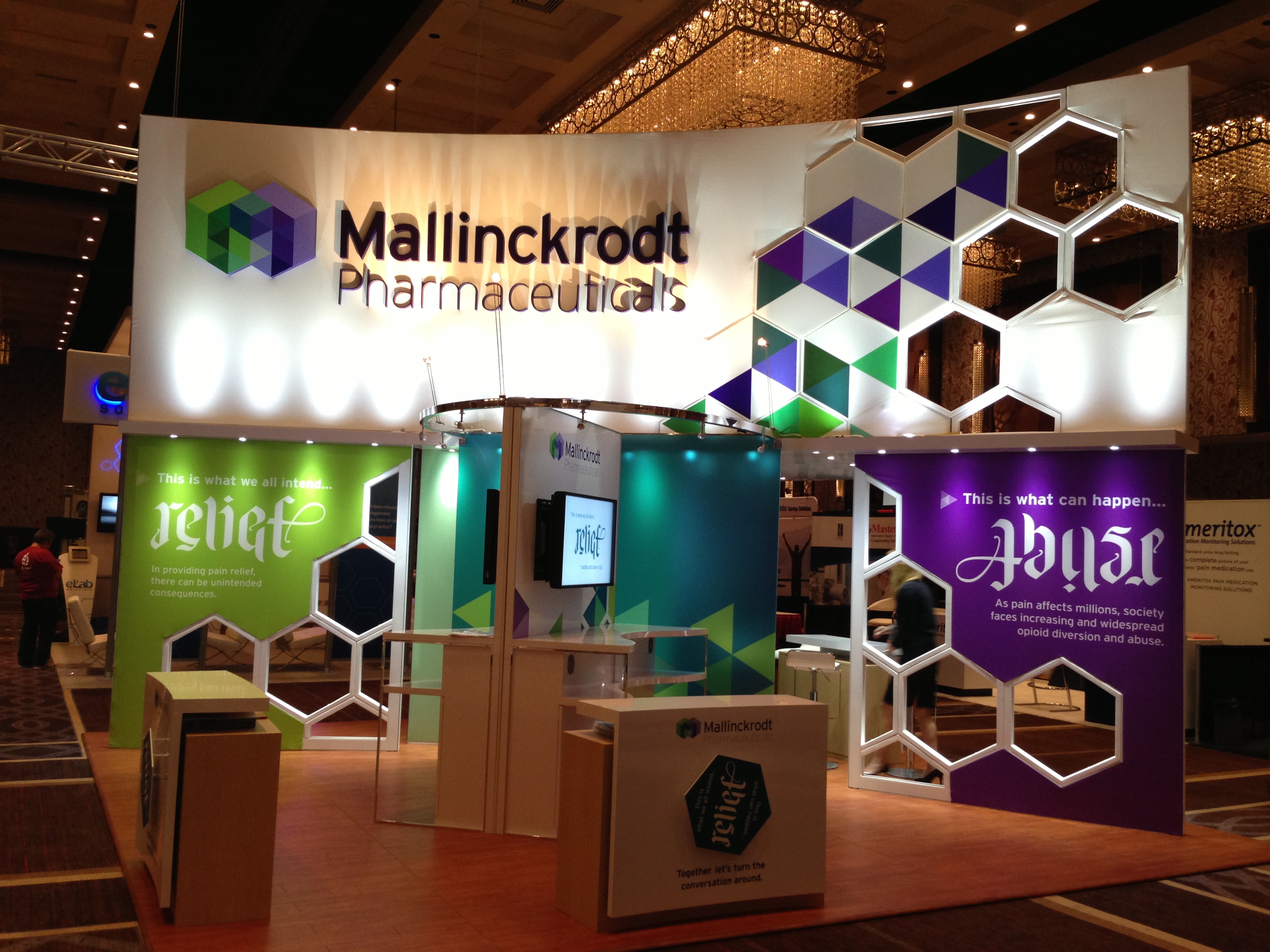 A trade show booth for Mallinckrodt Pharmaceuticals. The booth is decorated with a colorful geometric pattern in shades of purple green and blue. The company's logo is prominently displayed on the top left corner of the booth.<br /><br />The booth has a white background with the company's name written in bold black letters. Below the logo there is a large banner that reads "Relief" in a modern sans-serif font. On the right side of the image there are two smaller banners that read "Abuse" in purple and green letters. The booths are arranged in a semi-circular pattern with a large chandelier hanging from the ceiling in the background. The floor is made of wood and there are several people walking around the booth admiring the design.