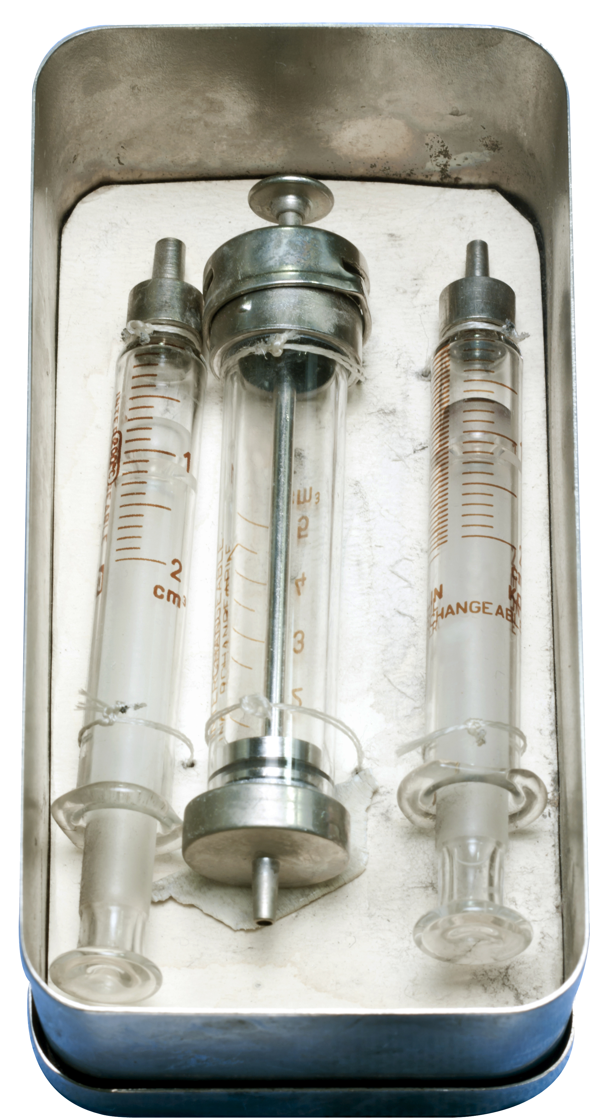 Three syringes in a metal tray. The tray is rectangular in shape and has a handle on top for easy carrying. The syringes are made of glass and have a cylindrical body with a needle at the end. The needle is marked with numbers and letters indicating the size of the syringe. There is also a label on the side of each syringe that reads "Changeable". The tray appears to be old and dirty with some dirt and grime visible on the surface.