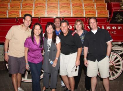 A group of seven people standing in front of a red fire truck. The truck has the words "Pilsner Inc." written on the side in white letters. The people are all smiling and appear to be posing for a photo. The woman in the center is wearing a purple blouse and is holding a water bottle. The man on the left is wearing khaki shorts and a beige t-shirt. The other seven people are wearing casual clothing. The background appears to be a brick building with a brick wall.