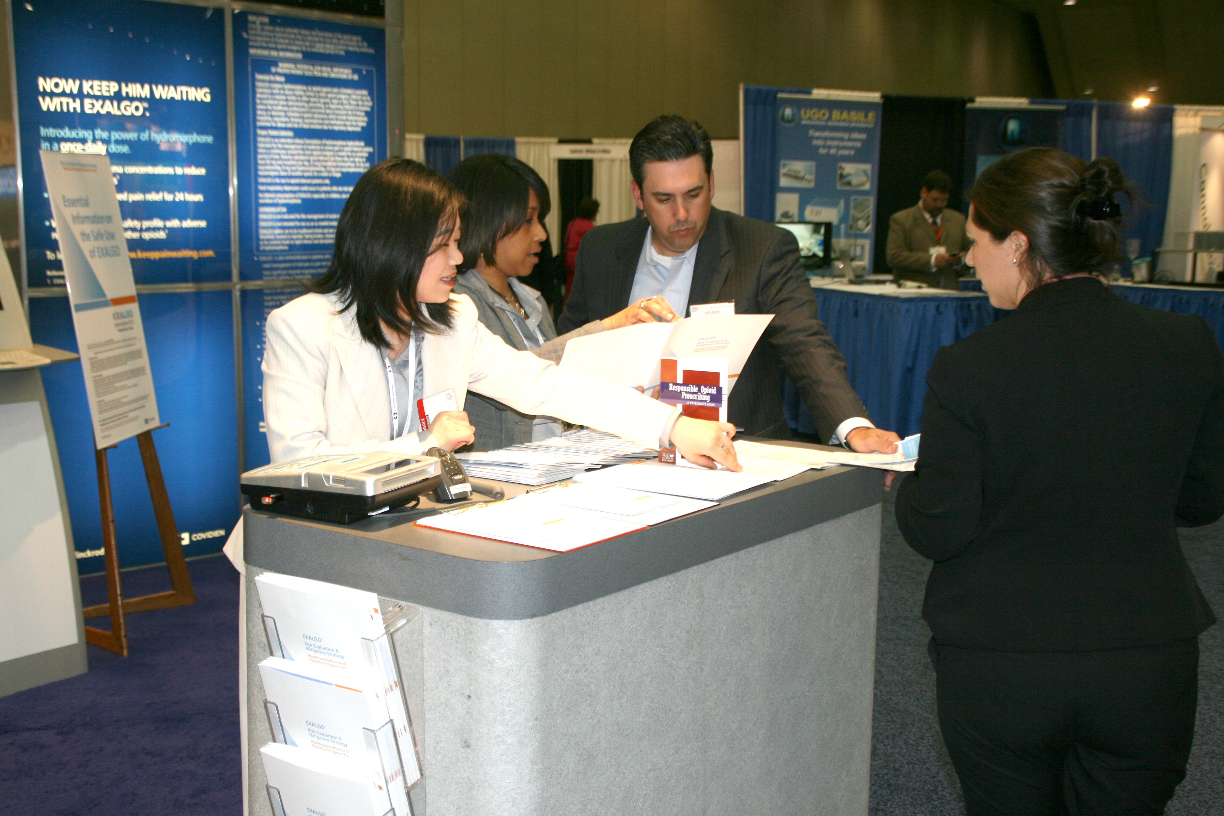 A group of four people at a trade show booth. There are three people in the image two women and one man standing behind a reception desk. The woman on the left is wearing a white blazer and is holding a stack of papers while the man in the middle is holding some papers and appears to be explaining something to the two women on the right. The man is also holding a piece of paper and is looking at it intently. In the background there are blue banners with white text and images as well as other booths and people. The booth is set up in a large room with a blue carpet and white walls.