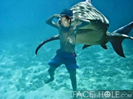 A young man swimming with a large shark in the ocean. The man is wearing blue shorts and is holding the shark with both hands. The shark is grey and black in color and is swimming towards the man. The water is clear and blue and there are small rocks visible at the bottom of the ocean floor. The image appears to be taken underwater as the man is in the center of the frame.