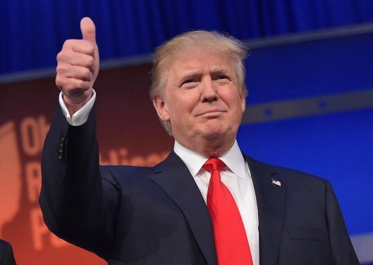 Former US President Donald Trump giving a thumbs up on a stage. He is wearing a dark suit and a red tie and is standing in front of a blue curtain with the words "Donald Trump" written on it. He appears to be giving a speech or making a point with his right hand. His facial expression is serious and he is looking directly at the camera.