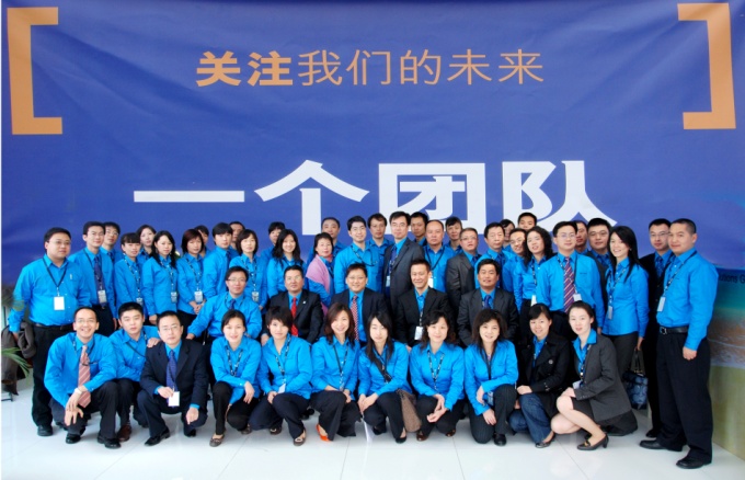 A group of people posing for a group photo in front of a large blue banner with Chinese characters written on it. The banner appears to be a banner for a company or organization as there is a logo of a building in the background. The people in the photo are all wearing blue shirts and are arranged in a semi-circle with some standing and some kneeling. They are all smiling and looking at the camera. The overall mood of the photo is professional and celebratory.