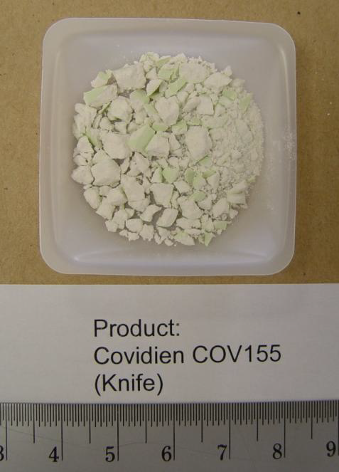 A small white rectangular container with a small amount of white powder inside. Its label reads "Product: Covidien COV155 (Knife)". The container is placed on a beige surface and there is a ruler next to it indicating that the powder is being measured.