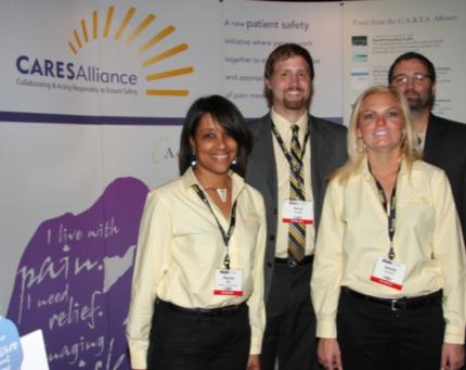 Four people standing in front of a banner that reads "CARESAlliance". The banner also has the logo of the organization and the words "A new patient safety initiative" written on it. The people in the image are two men and two women all wearing name tags. They are all smiling and appear to be posing for a photo. The background is a white wall with a purple banner on the left side.