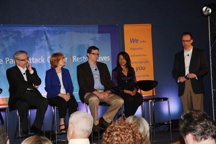 A group of five people sitting on a stage in front of a blue background with a banner that reads "We make diagnostics better for you". There are four people on the stage three men and two women all dressed in formal attire. The man on the left is wearing a black suit and glasses the man in the center is holding a microphone and the woman on the right is holding an orange banner with the words "Restore Lives" written on it. The audience is seated in rows of chairs facing the stage and attentively listening to the presentation.