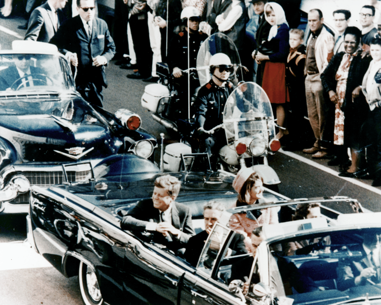 A black and white photograph of a group of people gathered on a street. In the center of the image there is a man sitting in the driver's seat of a black convertible car. He is wearing a suit and tie and appears to be speaking into a microphone. To his left there are two other cars one of which is a vintage car and the other is a newer one. On the right side of the photo a man in a suit is standing next to a motorcycle with a helmet on. Behind the motorcycle a crowd of people can be seen standing and watching the scene unfold. The people in the photo appear to be dressed in formal attire with some wearing hats and others wearing suits. The street is lined with buildings and there are trees in the background.