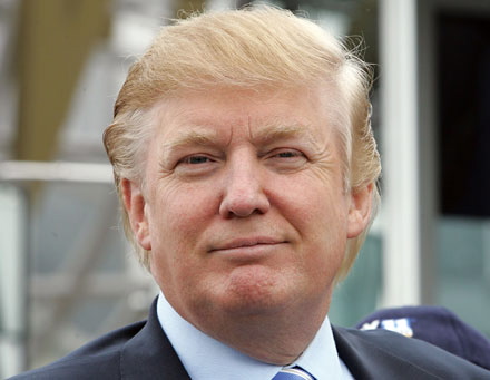 A close-up portrait of former US President Donald Trump. He is seen from the side looking directly at the camera with a serious expression on his face. He has blonde hair and is wearing a dark suit and a blue tie. The background is blurred but it appears to be an outdoor setting with a building visible in the distance.