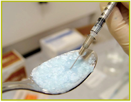 A close-up of a person's hand holding a syringe with a needle inserted into a spoon. The syringe is filled with a light blue substance which appears to be sugar or a similar substance. The spoon is silver and has a handle on one side. The background is blurred but it seems like the person is in a laboratory or a medical setting.