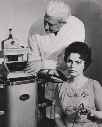 A black and white photograph of an elderly man and a young woman in a laboratory setting. The man is standing behind the woman who is sitting in front of a large machine. The woman is holding a small device in her hand and appears to be examining the machine with it. Both the man and the woman are wearing lab coats and appear to be focused on the task at hand. The machine is a scientific instrument with a cylindrical body and a handle on top. The background is plain and the overall mood of the image is serious and focused.