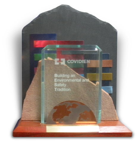 Of a glass award with a wooden base. The award is in the shape of an arch with a curved top and a flat base. Inside the arch there is a text that reads "COVIDIEN" and "Building an Environmental and Safety Tradition". The text is written in different colors including red blue green yellow and orange. The background of the award is black and there is an image of a globe in the bottom right corner.