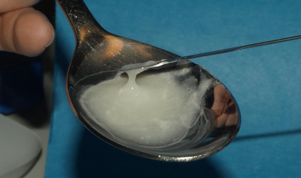 A hand holding a silver spoon with a small amount of white substance likely heroin or another drug on it. The spoon is resting on a blue surface and the person's hand is visible on the left side of the image. A small scoop is being used to scoop or stir the substance. The background is blurred but it seems like the focus is on the spoon and the substance.