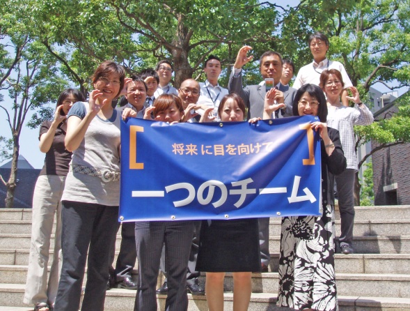 A group of people standing on a set of stairs in a park. They are holding a large blue banner with Japanese text on it. The banner appears to be a protest or rally as the text is written in both English and Japanese. The people in the group are smiling and making peace signs with their hands. Some of them are wearing casual clothes while others are dressed in formal attire. The background shows trees and buildings suggesting that the photo was taken in a city or urban area.