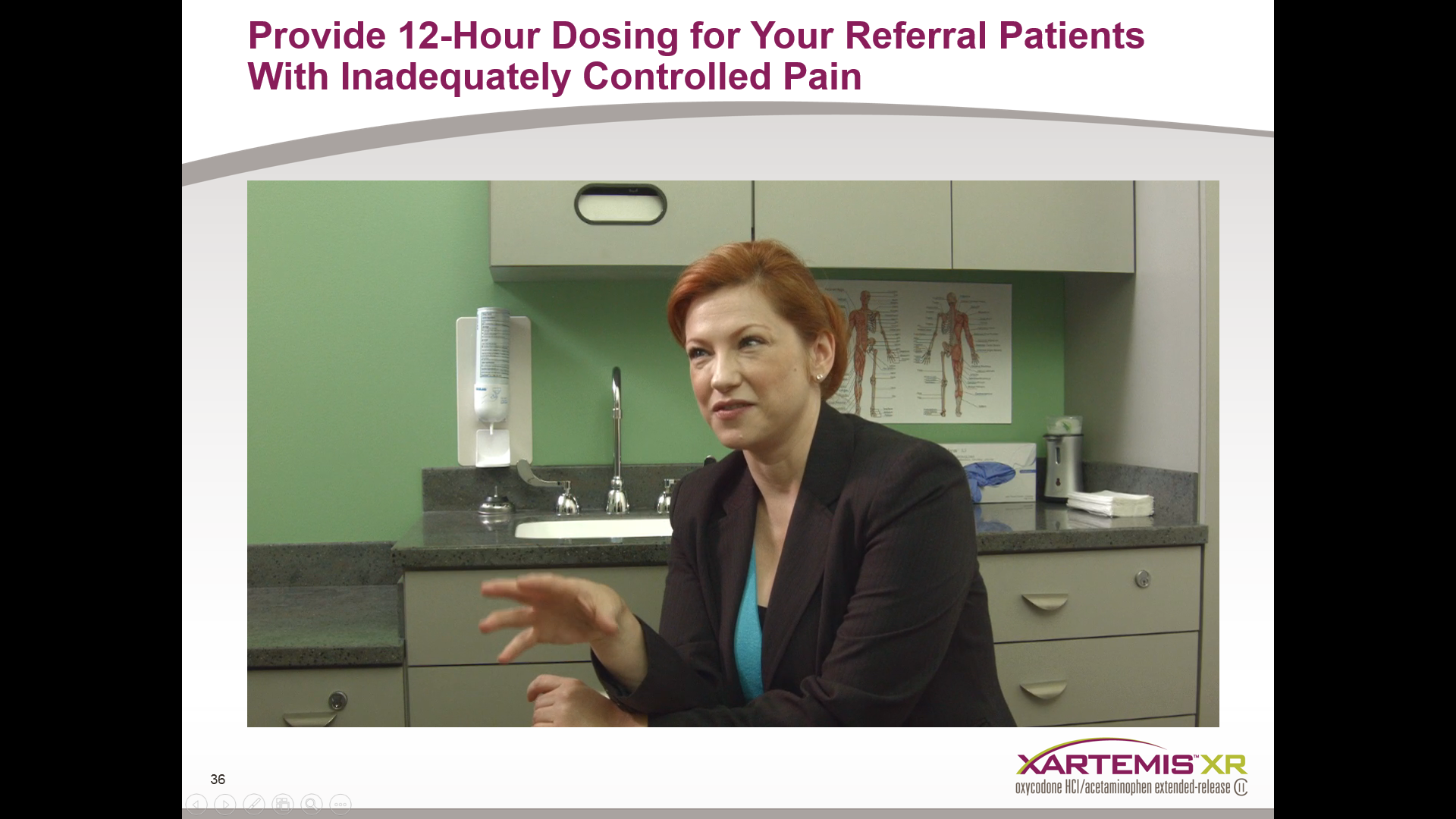 A woman sitting in a medical office with a green wall and a sink in the background. She is wearing a black blazer and appears to be in the middle of a conversation. On the wall behind her there is a poster with a diagram of the human body and text that reads "Provide 12-Hour Dosing for Your Referral Patients With Inadequately Controlled Pain". The woman is gesturing with her hands as if she is explaining something.