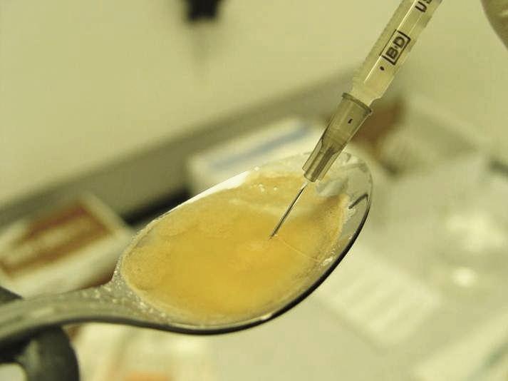 A close-up of a spoon with a syringe attached to it. The syringe is filled with a yellow liquid which appears to be a mixture of some kind. The spoon is resting on a white surface and there are some papers and other medical equipment visible in the background. The image is taken from a slightly elevated angle looking down on the spoon and the syringe.