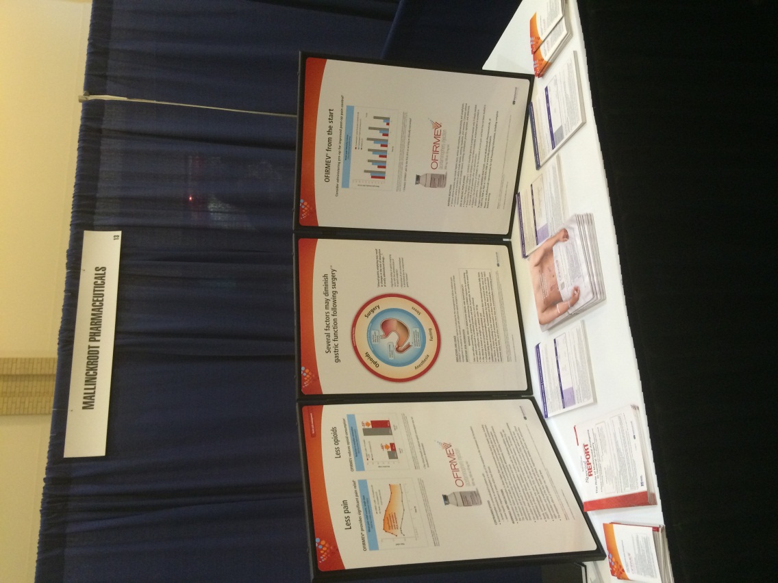 A table with a blue curtain in the background. On the table there are several brochures and pamphlets arranged in a grid-like pattern. The brochures are of different sizes and colors with some being white some being orange and some being blue. There is also a small white sign on the left side of the table that reads "Welcome to the conference". The table appears to be set up for a presentation or event.