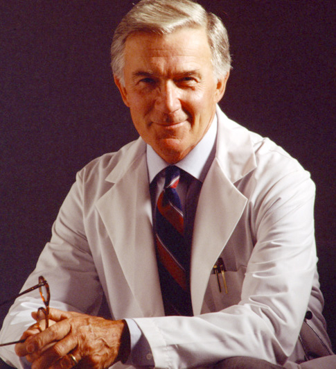 A portrait of a middle-aged man wearing a white lab coat and a red and blue striped tie. He is sitting in a chair with his legs crossed and is holding a pair of glasses in his left hand. He has a serious expression on his face and is looking directly at the camera. The background is dark making the man the focal point of the image. He appears to be in his late 60s or early 70s.