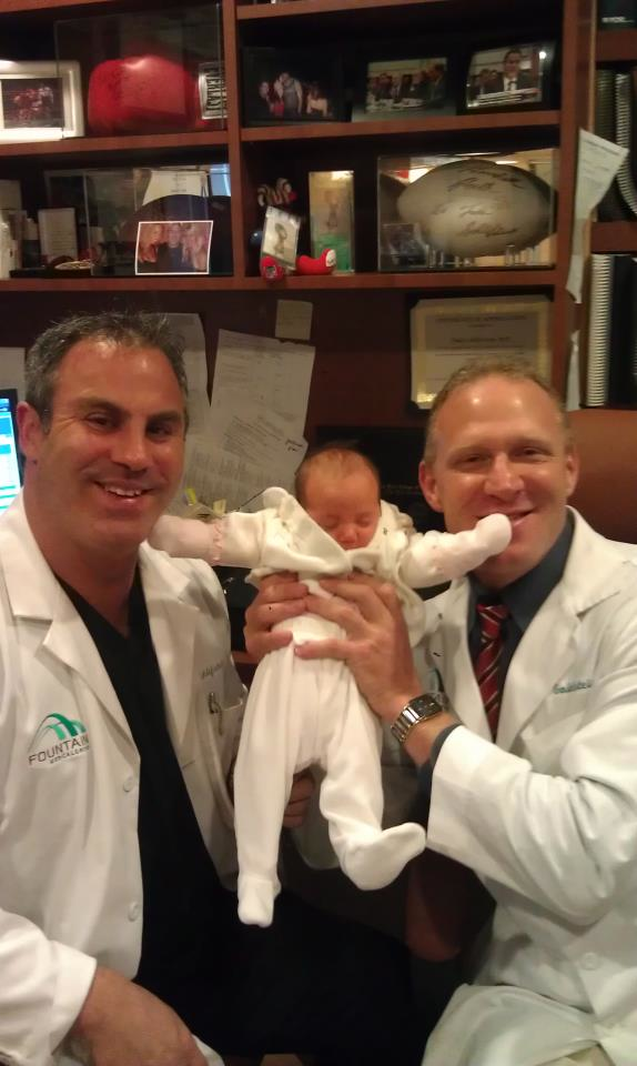 Two men in white lab coats sitting in an office. They are both smiling and holding a newborn baby in their arms. The baby is wearing a white onesie and is wrapped in a white blanket. The man on the left is holding the baby close to his chest and is looking at the camera with a smile on his face. On the right side of the image there is a desk with a computer monitor and other office supplies. In the background there are shelves with various items on them.