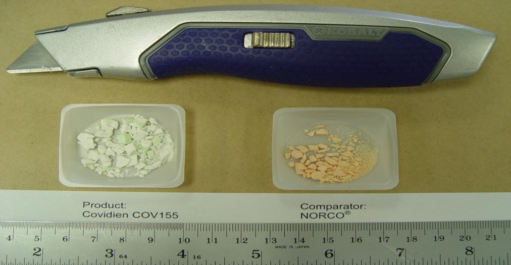 A blue and silver folding knife with a blue handle. The knife has a sharp blade and a pointed tip. Next to the knife there are two small plastic containers with white and green pills in them. The containers are labeled "Product: Covidien COV155" and "Comparator: Norco". The background is a beige surface.