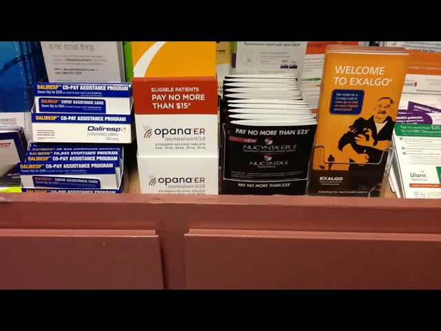 A shelf in a store with various items on it. On the left side of the shelf there are several boxes of Opana ER products including a sign that reads "Pay No More Than $15". On the right side there is an orange box with the words "Welcome to Exalgo" written on it and a black box with an image of a man holding a baby. In the center of the image there appears to be a stack of white boxes with the same text. The boxes are arranged neatly on the shelf. The background is blurred but it seems to be an indoor space with other items visible.