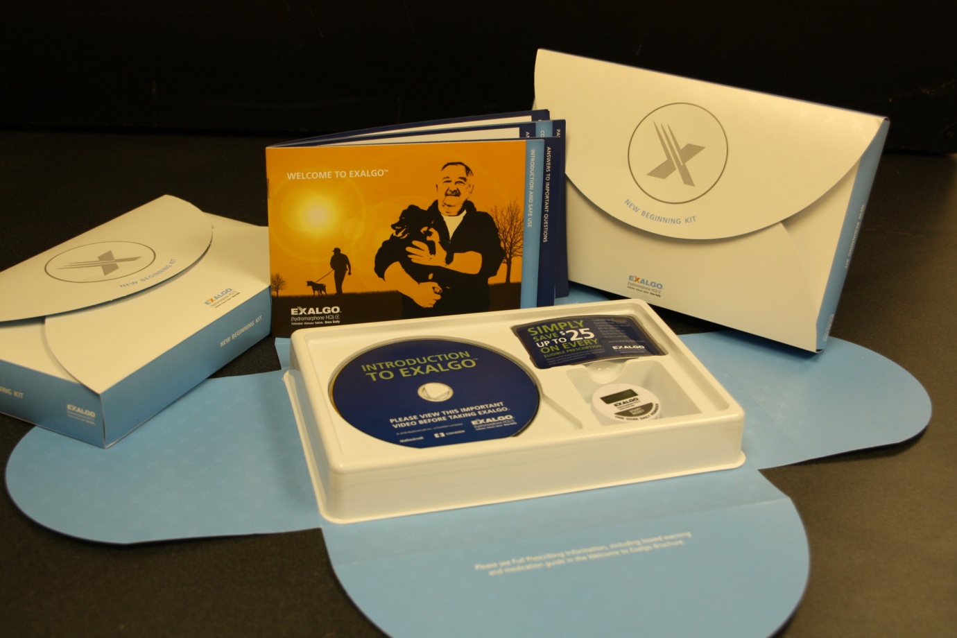 A collection of items arranged on a black surface. There are three items in the image - a white envelope with a blue label on the left side a blue CD case with a CD inside and a white box with a white cover. The CD case has a picture of a man holding a dog and the words "Welcome to Exalgo" written on it. The box also has a blue and white logo on the top right corner. The items appear to be new and unused with no visible damage or wear.