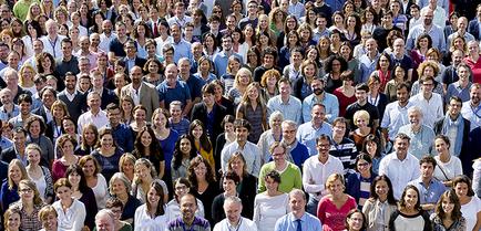 A large group of people standing in a grid-like pattern. The people are of different ages genders and ethnicity's and they are all smiling and looking at the camera. They appear to be diverse in age gender and ethnicity. Some people are wearing casual clothes while others are dressed in more formal attire. The background is blurred but it appears to be an outdoor setting with trees and buildings visible. The overall mood of the image is cheerful and happy.