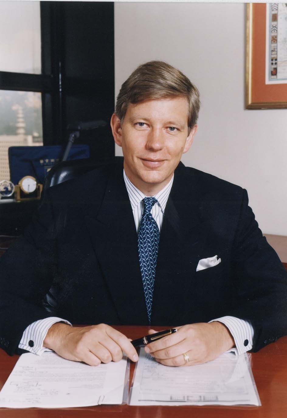 A portrait of a man sitting at a desk in an office. He is wearing a dark suit with a white shirt and a blue tie. He has short blonde hair and is smiling at the camera. The man is holding a pen in his right hand and appears to be signing a document. On the desk in front of him there are several papers and a clock. The background shows a window with a view of a city skyline.