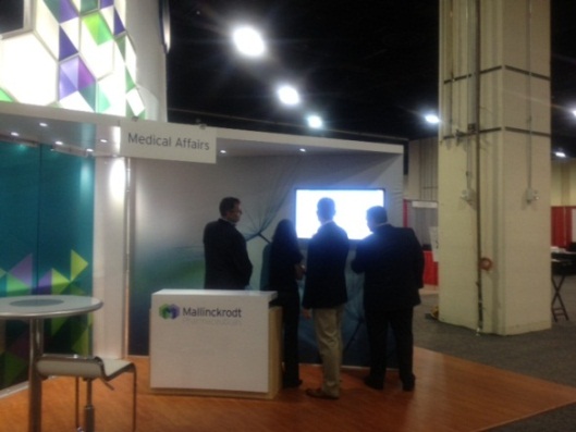 A medical affairs booth at a trade show. The booth is white with a colorful geometric pattern on the left side and a sign that reads "Medical Affairs". There is a small table in front of the booth and a few people standing around it. On the right side of the image there is a large screen displaying information about the booth. The floor is made of wood and there are other booths and tables visible in the background.