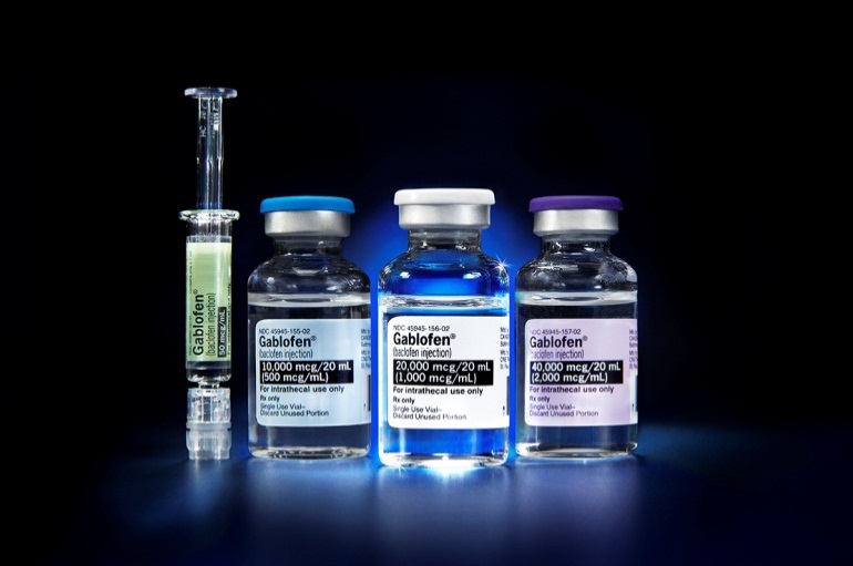 Three vials of different sizes and shapes arranged in a row on a dark blue background. The vials are of different colors - one is green one is blue and one is purple. Each vial has a white label with the name "Gabrielle" written on it. Next to the vials there is a syringe with a green liquid inside. The syringe appears to be filled with a clear liquid. The background is completely black making the colors of the vial and syringe stand out.