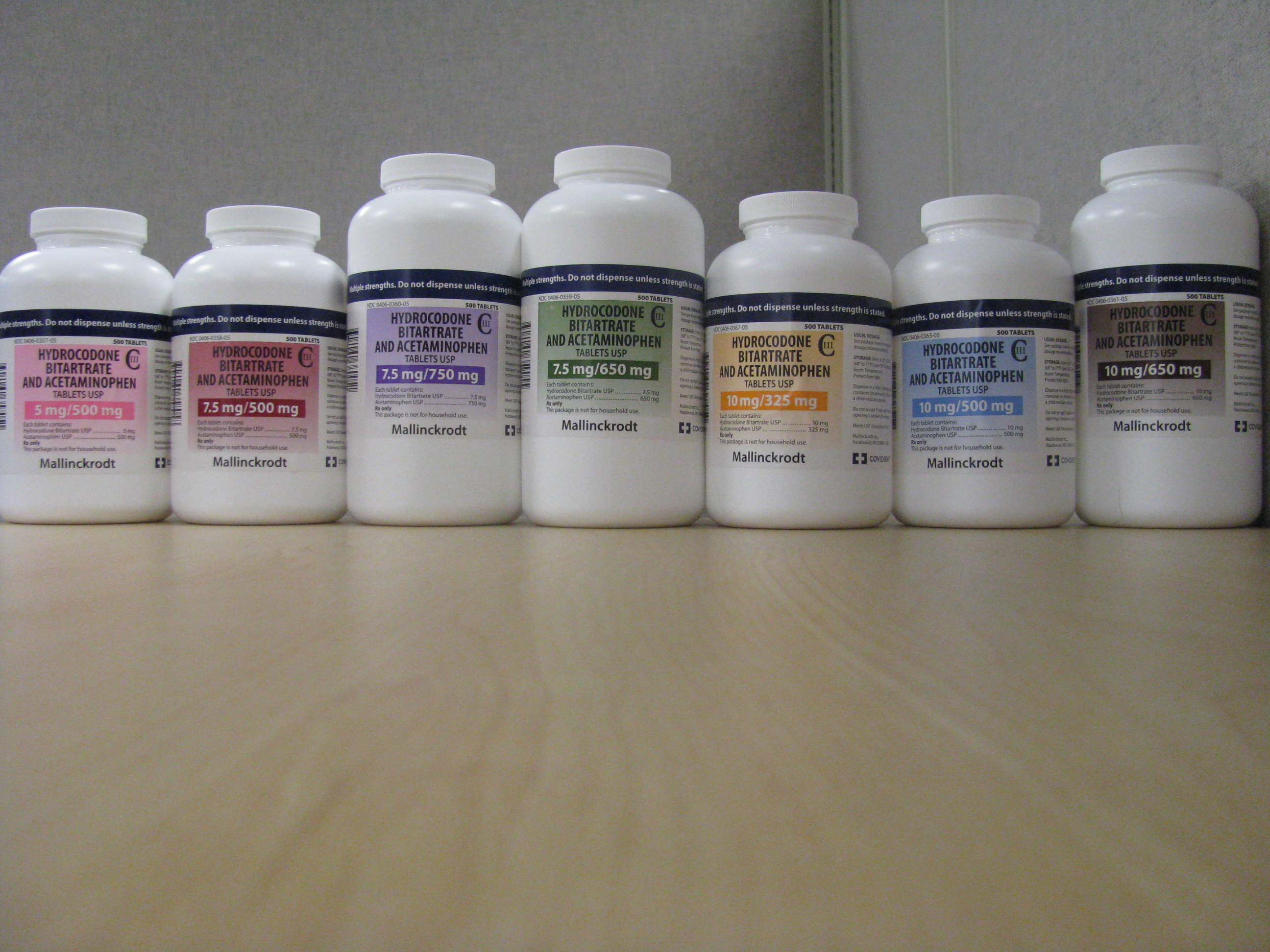 A row of nine white plastic bottles arranged in a line on a wooden surface. The bottles are of different sizes and shapes with the largest bottle at the front and the smallest at the back. Each bottle has a blue label with white text and a red and white logo. The labels appear to be for different types of supplements or supplements. The background is a plain white wall.