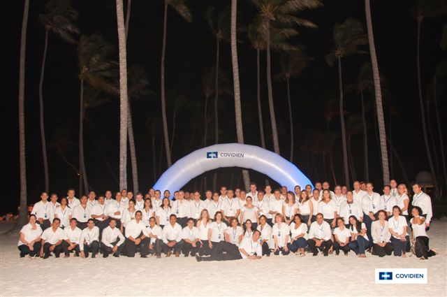 A group of people posing for a group photo on a beach at night. The group is arranged in a semi-circle with a large blue inflatable arch in the center. The arch has the word "COVIDIEN" written on it in white letters. The people are all wearing white shirts and black pants and they are all smiling and looking at the camera. The beach is lined with palm trees and the sky is dark suggesting that it is nighttime. The overall mood of the photo is celebratory and happy.